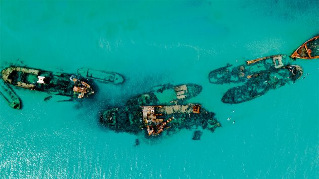 Aerial shot of clear water with a curved line of rusty shipwrecks.