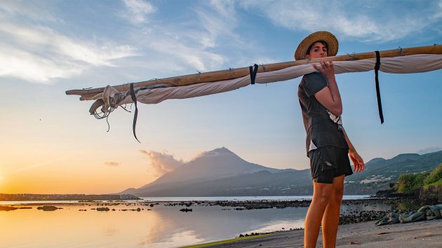 A man carries a gaff pole on his shoulder. The beach and mountain are behind with the setting sun.