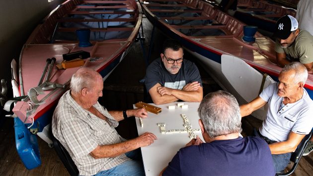 A group play dominoes on a table in a boatshed.